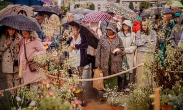Visitors holding umbrellas or in raincoats with hoods up stand at the other side of a rope that separates them from a garden at the show