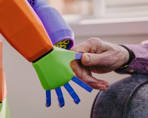 An elderly man's hand holds a multicoloured robot hand belonging to Abi the robot