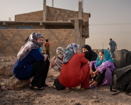 Women and girls squat by their suitcases on a dirt road outside a house