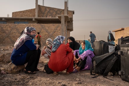 Women and girls squat by their suitcases on a dirt road outside a house