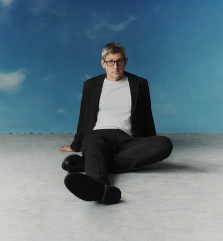 Louis Theroux wearing a black suit, a white T-shirt, black shoes and black-rimmed glasses, with his right leg on the floor in front of him and his left leg folded underneath it, his hands on the floor behind him, sitting on a grey floor with the sky as the background.