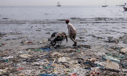 A man pushes a wheelbarrow full of plastic waste along a beach with more waste in the foreground