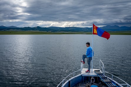 Bir adam göldeki bir teknenin önündeki direğe asılı bayrağın yanında duruyor