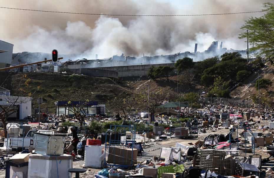 A factory burns in the background while empty boxes litter the road from looted goods being removed.