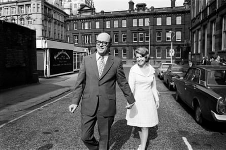 black and white photo of Anne Reid in a white knee-length dress holding hands with Peter Eckersley in a suit as they walk down a street