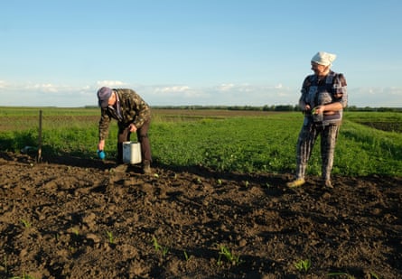 A woman stands in a field watching a man bent over carrying a small pot and a larger tub