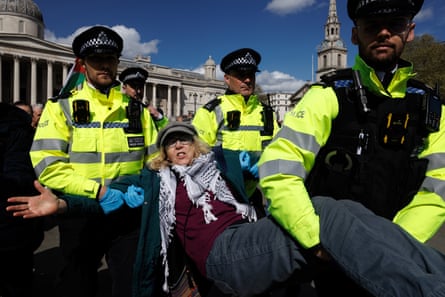 Police detain a person wearing a keffiyeh by carrying them by their arms and legs