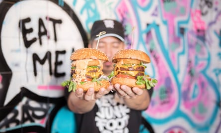 Employee holds up two vegan burgers in front of a colourful mural that says Eat Me! at Vegan Junk Food Bar in Amsterdam, the Netherlands.