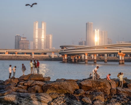 Local people stand on rocks by the sea with Mumbai’s coastal road in the distance with the high-rise blocks of the city behind.