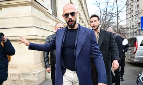 Andrew Tate in a T-shirt and blazer, wearing sunglasses and holding up his right hand, walks down a street in Bucharest in the middle of speaking