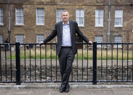 David Gauke stands against black iron railings in Westminster, with stone buildings in the background. He wears a dark grey suit with the jacket open over an open-necked white shirt. He has grey hair.