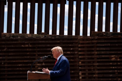 a man in a suit speaks into a microphone next to a border fence