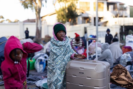Two girls wearing hats, coats and wrapped in a blanket stand next to a large suitcase with other people in the background sleeping on the ground outside