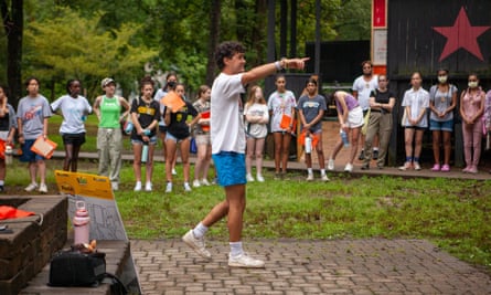 A large circle of students of many races, all wearing T-shirts and shorts, stand on a grassy lawn, facing a young man in the middle - who could be one of them or perhaps a young adult, such as a camp leader - wearing a white T-shirt and bright blue shorts, pointing with his right hand and smiling as he speaks.