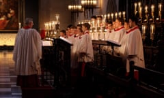 A choir master and two rows of boys from King’s College Cambridge choir, all in robes, in front of tall lit candles
