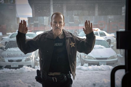 Bob Odenkirk dressed as a sheriff seen with his hands up through a window pane with a snowy urban landscape behind him
