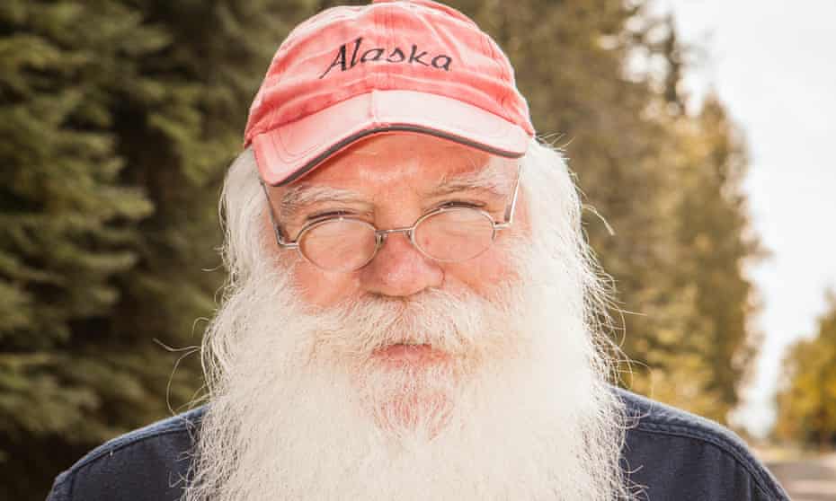 Portrait of a man with a long white beard and round, wire-rimmed glasses wearing a red ball cap with Alaska written on it.