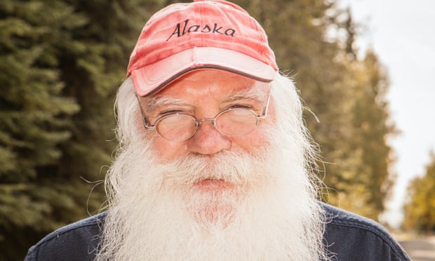 Portrait of a man with a long white beard and round, wire-rimmed glasses wearing a red ball cap with Alaska written on it.