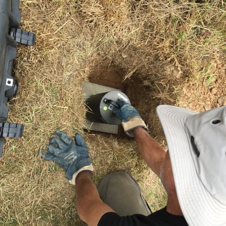 A man in a white hat, with safety gloves, puts a metal box of some description into a hole in the ground