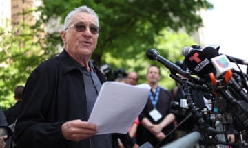 Older white man wearing sunglasses holds printouts and speaks into group of microphones, with trees behind him.