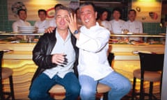 Nobuyuki Matsuhisa in a chef's jacket, with his arm around Robert De Niro, wearing a leather jacket, puts his hand up to De Niro's face as they are seated on high chairs in front of a sushi bar