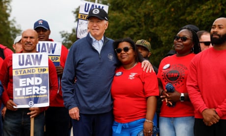An older white man wearing a dark blue baseball cap and dark blue fleece, stands close with a younger Black woman wearing sunglasses and a red T-shirt, his arm across her shoulders. Both are smiling, and they stand amid a crowd of other people wearing red T-shirts and holding signs that says 'UAW Stand Up End Tiers.'