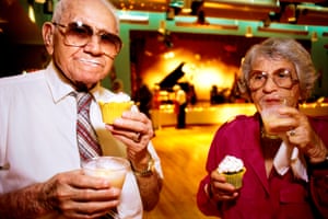 an older man and woman in a function room holding drinks glasses and cupcakes