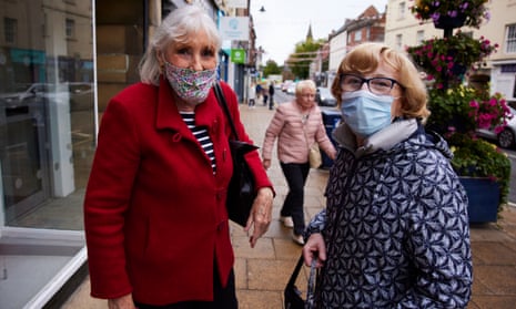 Two women wearing face masks and coats stand together in a high street