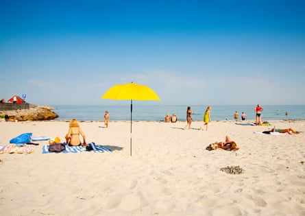 A woman sits on a striped towel next to a large yellow beach umbrella on a sandy beach with calm sea in the distance and other beachgoers