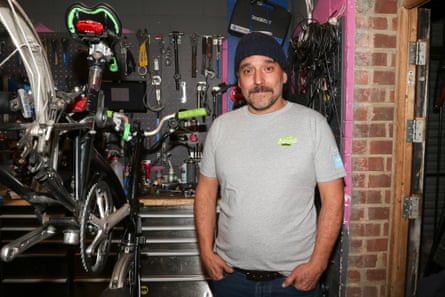 Tamas Polocz stands inside his bike shop with his hands in his jeans pockets. He is 49 with a short dark beard and brown eyes, and wears a black beanie hat and grey T-shirt. There is a bicycle on a rack to his side and lots of tools hanging on the wall behind him.