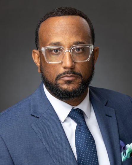 Headshot of a bearded man wearing a blue suit and tie and clear-rimmed glasses