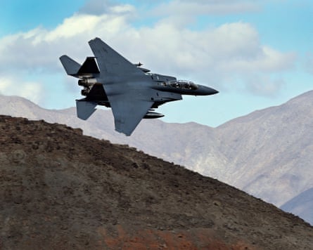 An F-15E Strike Eagle flies over a landscape with mountains and clouds in the background.