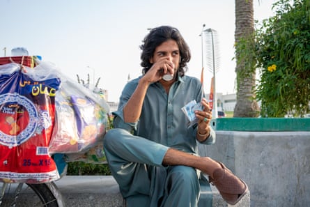 مردی در دوحه قطر در حال نوشیدن چای از یک فنجان کاغذی در کنار اسکله نشسته است.  او در هر انگشت یک حلقه با رنگ متفاوت دارد.
