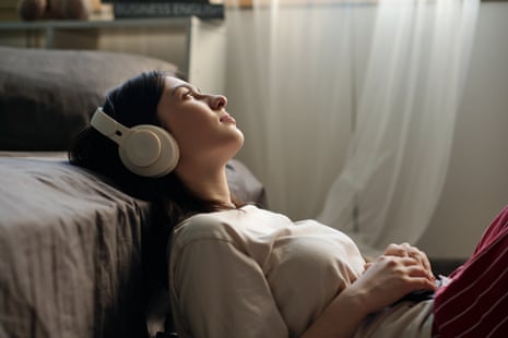 Caucasian young woman lying on floor beside bed listening to music with over ear headphones, gazing upward with relaxed expression, hands resting on stomach, bedroom setting visible