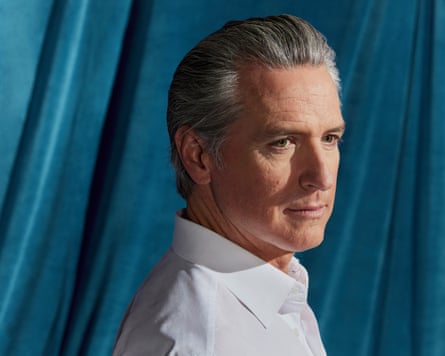 A head and shoulders shot of Gavin Newsom standing side-on to the camera, looking just to the right of it, wearing a white shirt and no tie, against a turquoise velvet background