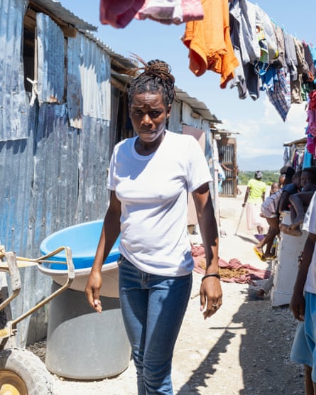 A woman in jeans and a tshirt stands in an alley next to a tin shelter with washing on a line above her