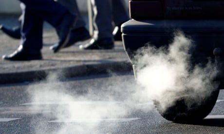 An exhaust pipe of a car on a street with legs of people walking past in view in the background