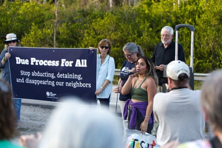 a woman speaks into a microphone next to people holding a banner that reads ‘due process for all’