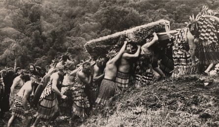 A large band of Māori men in traditional dress carry a coffin up a steep and rugged hill