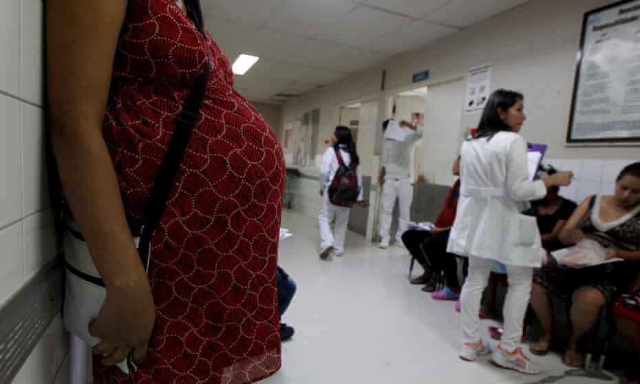 A pregnant woman in a red dress leans against a white clinic corridor wall; only her torso is visible.  Along the right hand side of the corridor are other women waiting on chairs, and white-coated clinic staff.