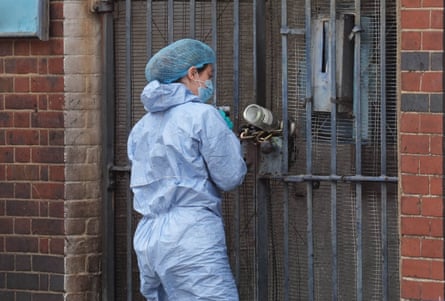 The investigator wearing light blue overalls, a hair-net and a face mask stands close to the entrance, where a bottle appears to be stuck between railings