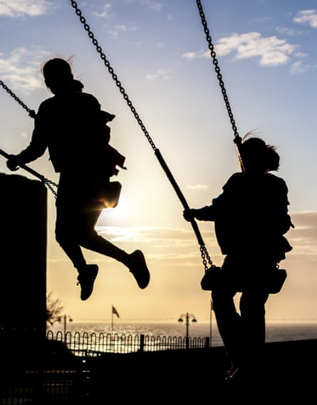 Silhouette of two female children playing on swings against a blue sky with the sun beginning to set
