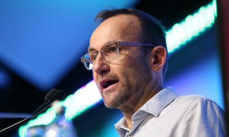 The Greens leader Adam Bandt addresses the Queensland Media Club in Brisbane, he is standing at a podium speaking into a microphone with bright green lights behind him