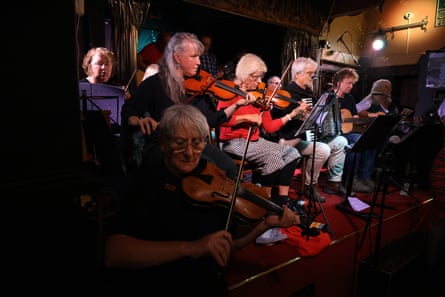 Violin, guitar and accordion players illuminated on the stage. They appear to mostly be older, grey-haired people.