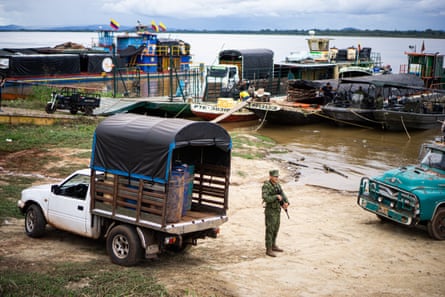 A man with a gun on the shore next to mining dredges, rafts with thatched roofs and mining equipment on them