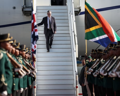 A man in a suit walk down the steps of a plane towards a guard of honour