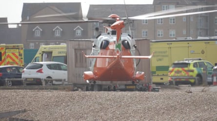 An orange air ambulance helicopter lands on the stony beach. Police cars and ambulances are parked in the background.