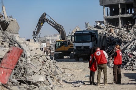 Three ICRC staff confer as an excavator behind them digs through piles of rubble