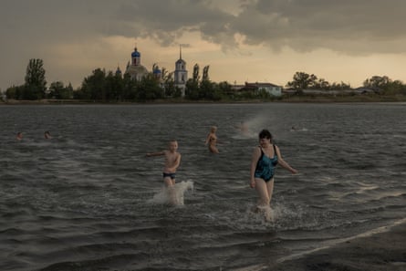 People swim in a lake and emerge from the water on a hot summer day; there is a church on the far bank and the sky is cloudy above.