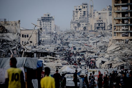 People walk through a street full of tents; half-destroyed buildings can be seen in the background